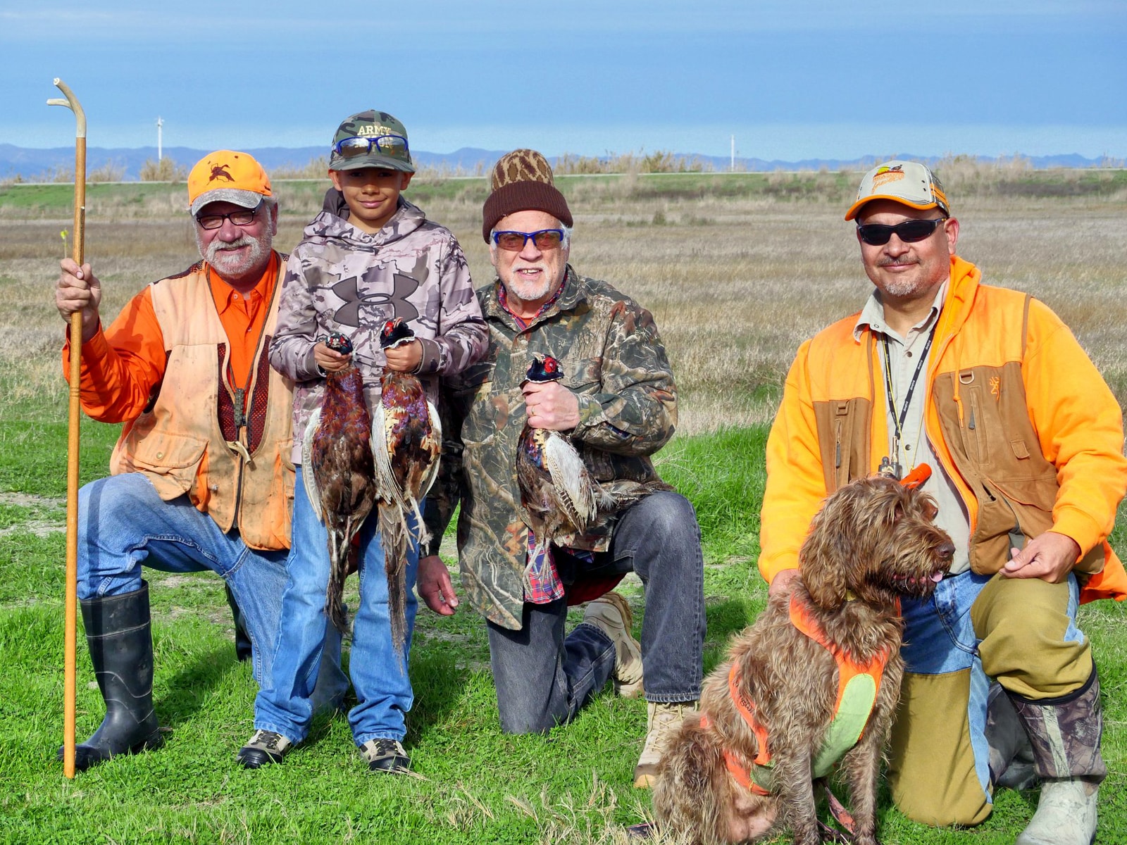 Group of four hunters including a young boy in an Army cap holding harvested pheasants with a Wirehaired Pointing Griffon in a sunny green field