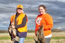 Man in blaze orange vest and sunglasses holding a harvested pheasant and shotgun in a field under overcast skies