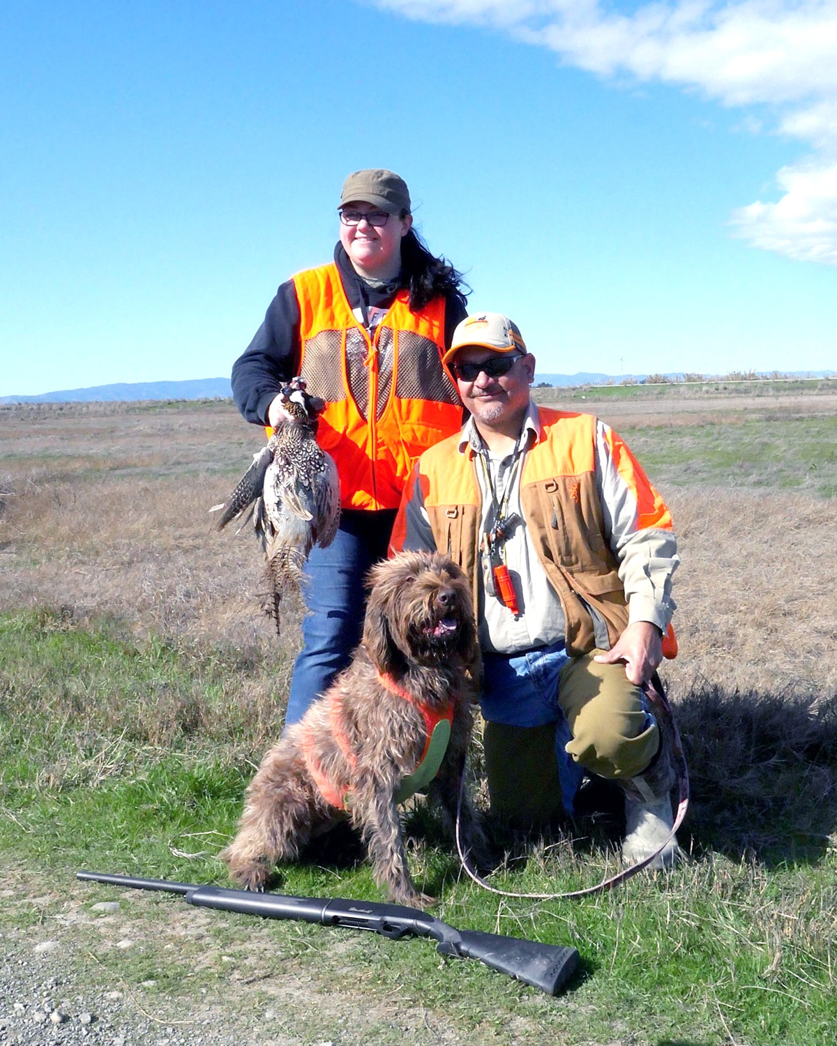 Woman hunter holding a pheasant and man kneeling with a Wirehaired Pointing Griffon dog and shotgun on the ground in a field