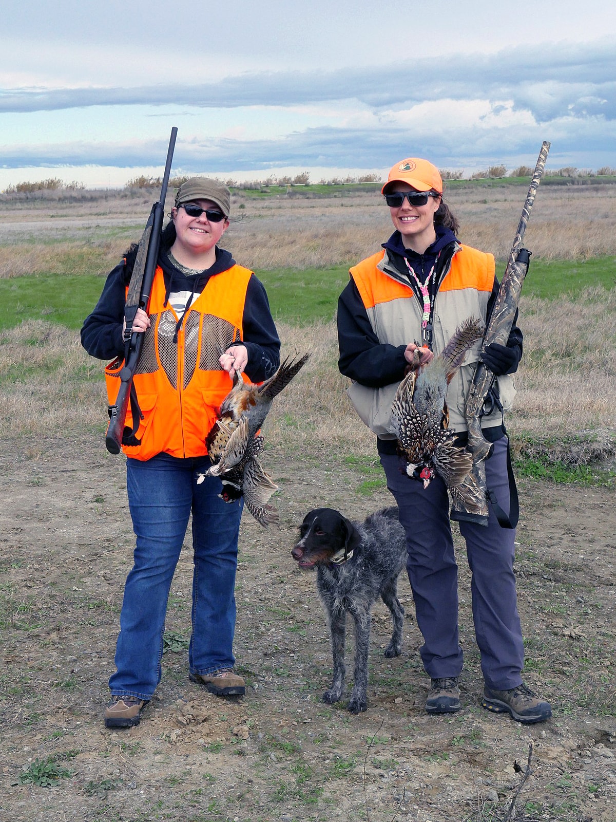 Two women hunters in blaze orange vests each holding harvested pheasants with a Wirehaired Pointing Griffon between them