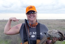 Woman in blaze orange cap and grey shirt flexing her arm while holding a harvested pheasant in an open field