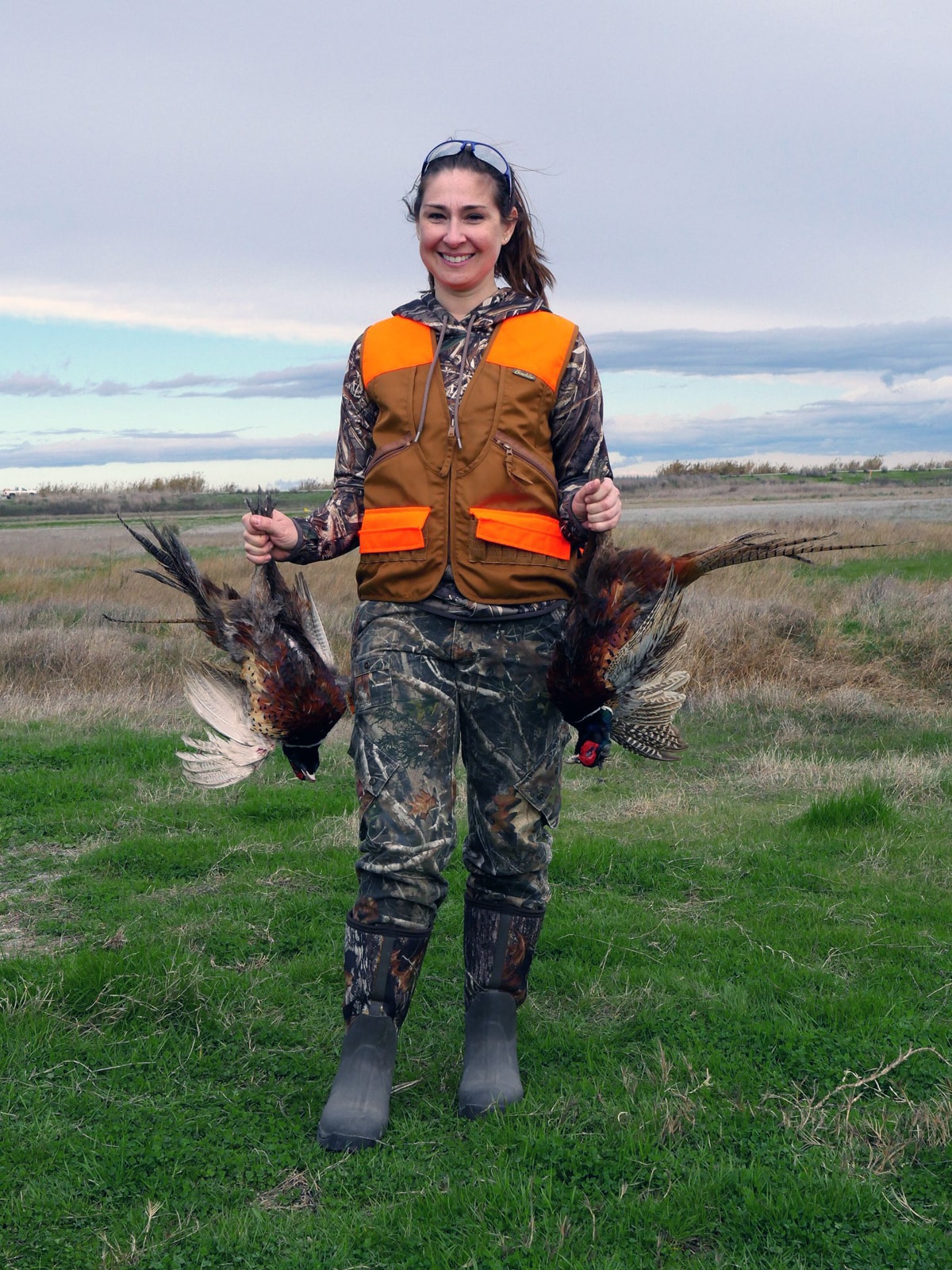 Woman hunter in blaze orange beanie and camo vest holding a harvested rooster pheasant and shotgun in an open field