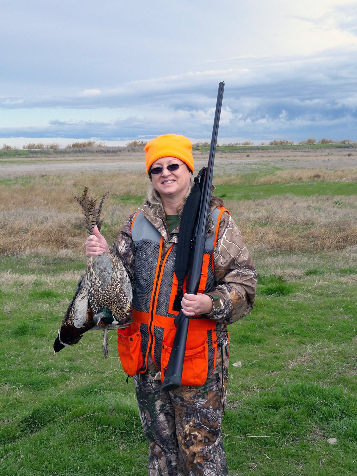 Young woman hunter in blaze orange vest holding a shotgun and harvested pheasant in a field under cloudy skies