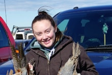 Smiling young woman in a brown jacket holding two harvested pheasants in a parking area with vehicles in the background