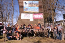 Large group of youth hunters, mentors, and chapter members posing in front of a barn with sponsor banners and their pheasant harvest at the 2022 Youth Mentor Hunt