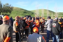 Group of six hunters in blaze orange including youth participants posing with two hunting dogs in a green rolling-hills field at the 2022 Youth Mentor Hunt