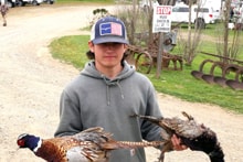 Young boy in a blaze orange vest and yellow cap holding two harvested pheasants on a gravel road at the 2022 Youth Mentor Hunt