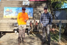 Two hunters in blaze orange and casual gear posing in front of the Pheasants Forever chapter sign with pheasants hanging on the bench at the 2023 Veterans and First Responders Hunt