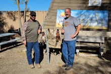 Two hunters standing together in front of the Pheasants Forever Yolo County chapter sign with harvested pheasants on the bench rack at the 2023 Veterans and First Responders Hunt