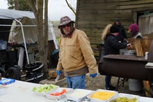Man in a tan jacket and blue gloves preparing food at an outdoor cookout table with condiments and toppings at the 2023 Youth Mentor Hunt