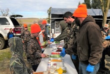Hunters in blaze orange lined up at an outdoor food table being served at the post-hunt cookout at the 2023 Youth Mentor Hunt