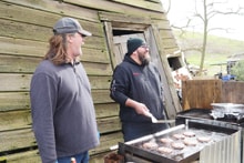 Two men grilling burgers on a large outdoor grill beside a weathered barn at the 2023 Youth Mentor Hunt