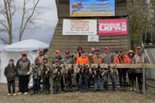 Group of youth hunters and mentors in blaze orange posing in front of a barn with sponsor banners and their pheasant harvest at the 2023 Youth Mentor Hunt