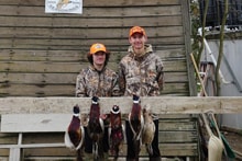 Two youth hunters in blaze orange and camo standing in front of a barn displaying their harvested pheasants at the 2023 Youth Mentor Hunt