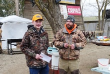 Young hunter in blaze orange cap holding a certificate while standing with a chapter member at the 2023 Youth Mentor Hunt prize and awards presentation