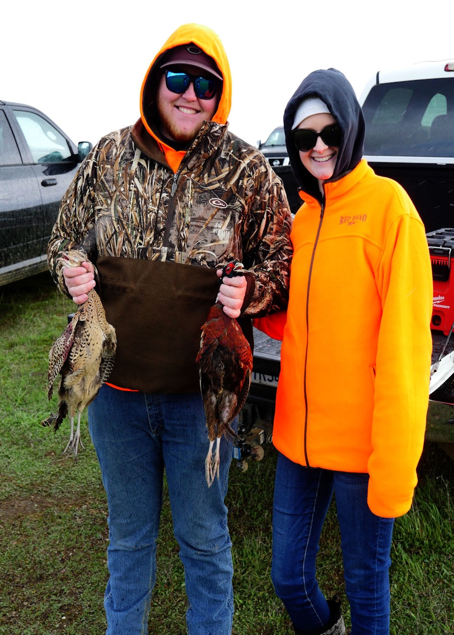 Man in camo hoodie and blaze orange and woman in a Red Head blaze orange jacket smiling together at a truck tailgate, each holding harvested pheasants on a rainy day at the 2024 R3 Upland Bird Hunt