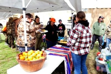 Hunters in camo and blaze orange serving themselves food at an outdoor tent buffet table with a basket of lemons at the 2024 Women's Upland Bird Hunt post-hunt cookout