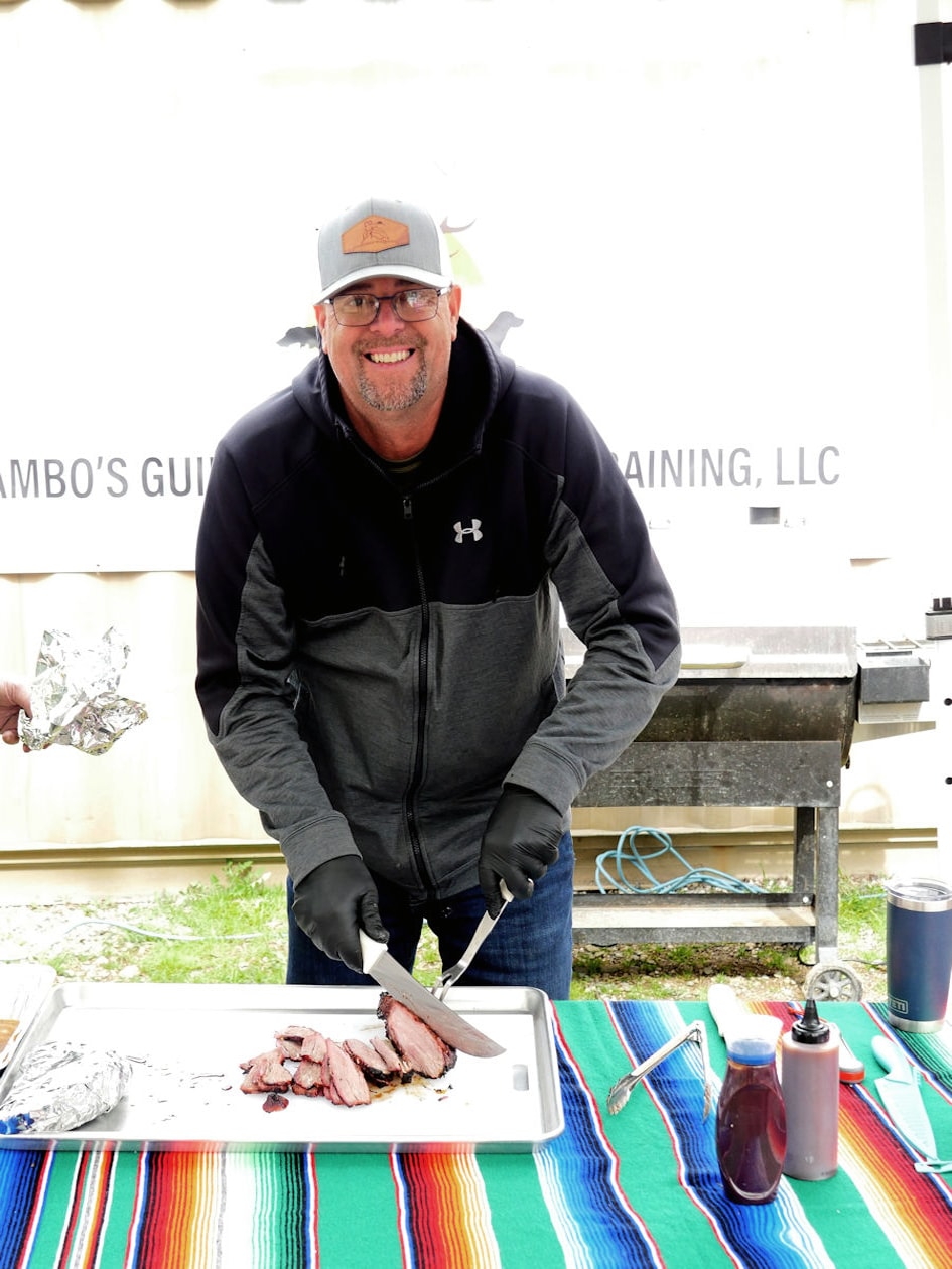 Smiling man in a grey hoodie and Rambo's Guide and Training cap carving smoked brisket on a cutting board at a colorful serape-covered table at the 2024 Women's Upland Bird Hunt post-hunt cookout