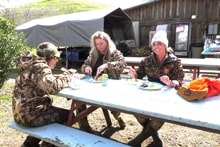 Three women in camo gear sitting together at a blue picnic table outside the lodge at the 2024 Women's Upland Bird Hunt