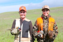 Two women hunters — one in a tan shirt and orange cap, one in a blaze orange vest and camo — each holding harvested quail in a green rolling-hills field at the 2024 Women's Upland Bird Hunt