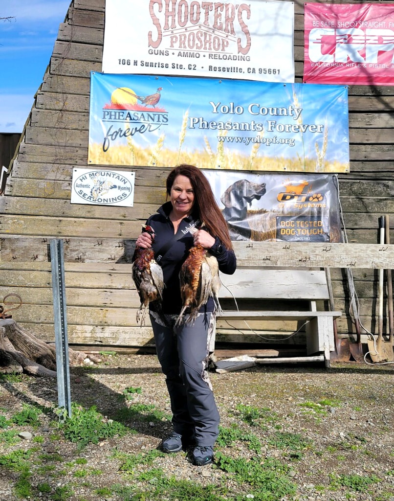 Woman in a black jacket smiling and holding two harvested pheasants in front of the Yolo County Pheasants Forever, Shooter's Pro Shop, CRPA, and DT Systems sponsor banners at the 2024 Women's Upland Bird Hunt