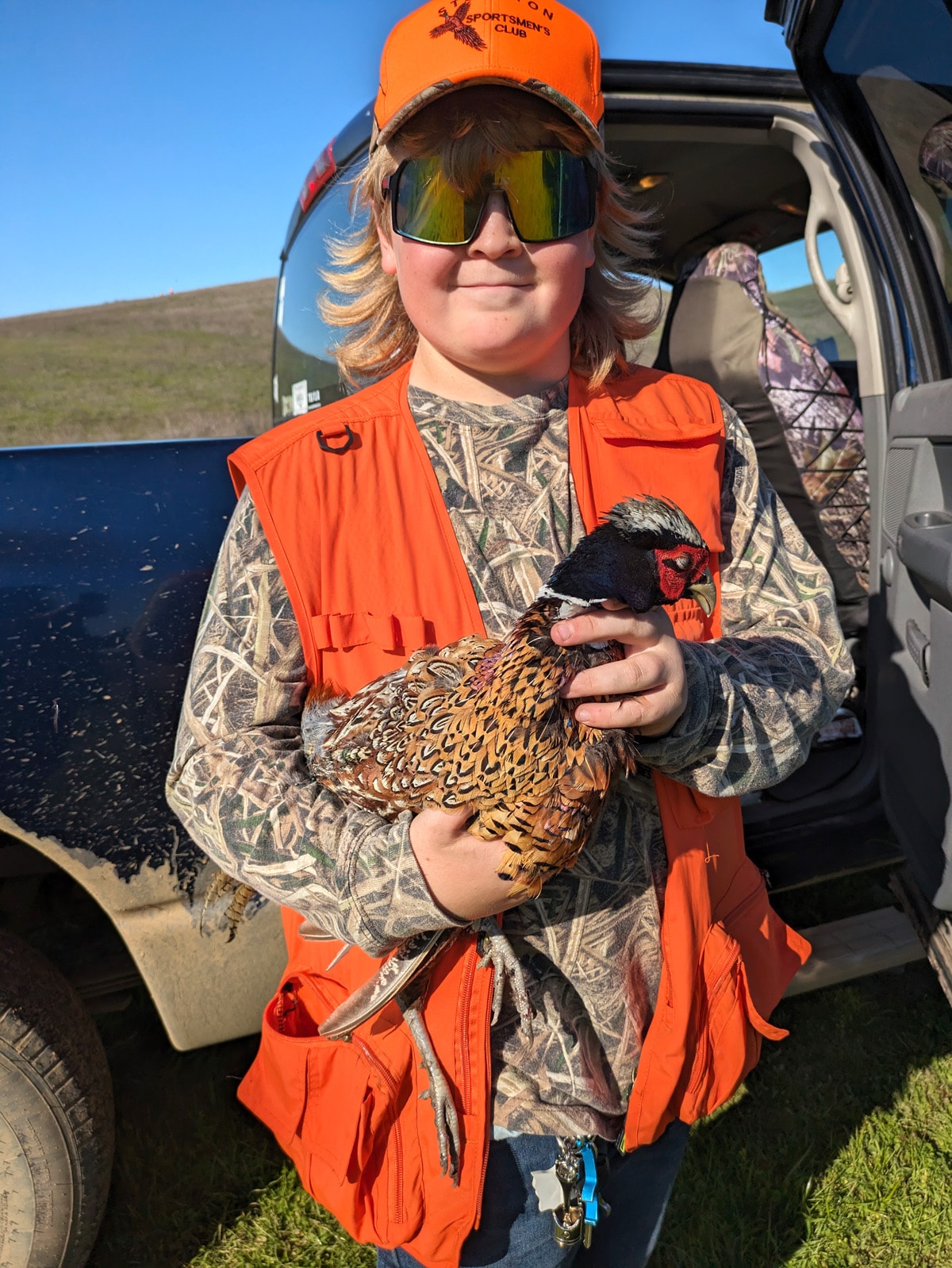 Boy in a blaze orange vest holding a harvested pheasant and a black shotgun against a green hillside at the 2024 Youth Mentor Hunt