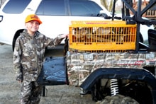 Young boy in a blaze orange vest holding a harvested pheasant and a shotgun with rolling green hills and blue sky behind him at the 2024 Youth Mentor Hunt