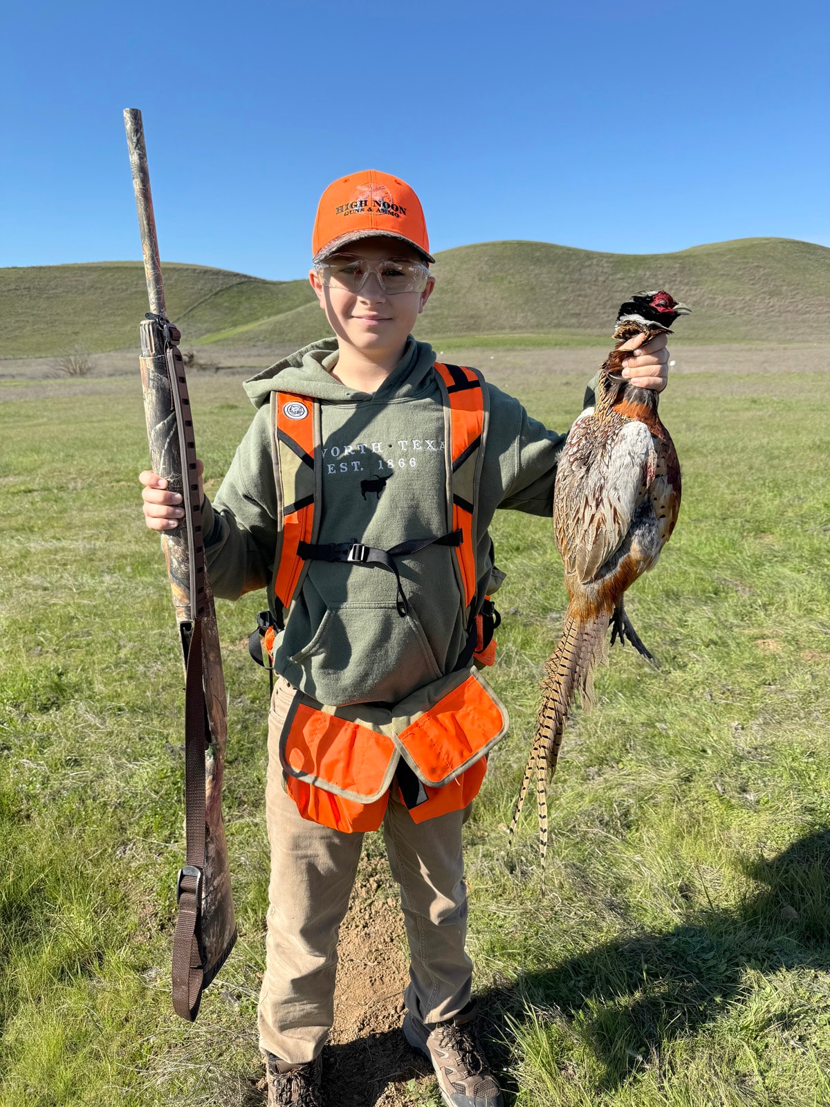 Boy in a blaze orange High Noon Guns cap and orange hunting vest holding a shotgun and a harvested pheasant in a green rolling-hills field at the 2024 Youth Mentor Hunt