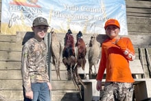 Two boys in camo and blaze orange standing in front of the Pheasants Forever Yolo County banner with harvested pheasants hanging on the rack at the 2024 Youth Mentor Hunt