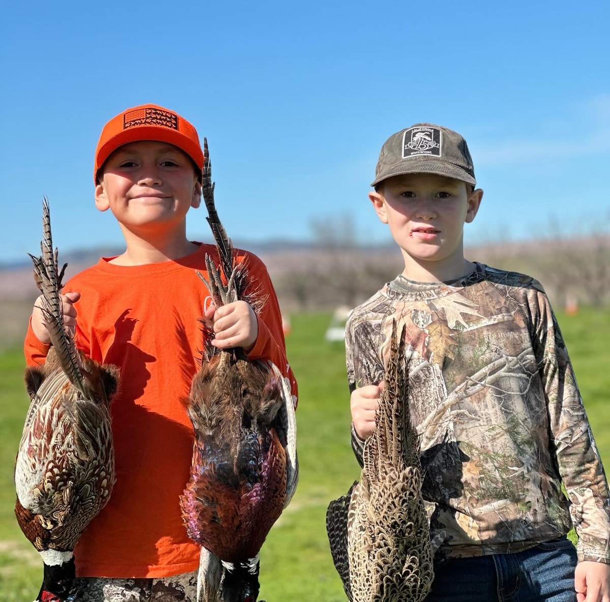 Two boys — one in an orange shirt and blaze cap, one in camo — each holding harvested pheasants in a green field under a blue sky at the 2024 Youth Mentor Hunt