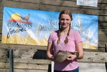 Youth hunter in blaze orange vest and camo holding a harvested pheasant with rolling green hills in the background at the 2024 Youth Mentor Hunt