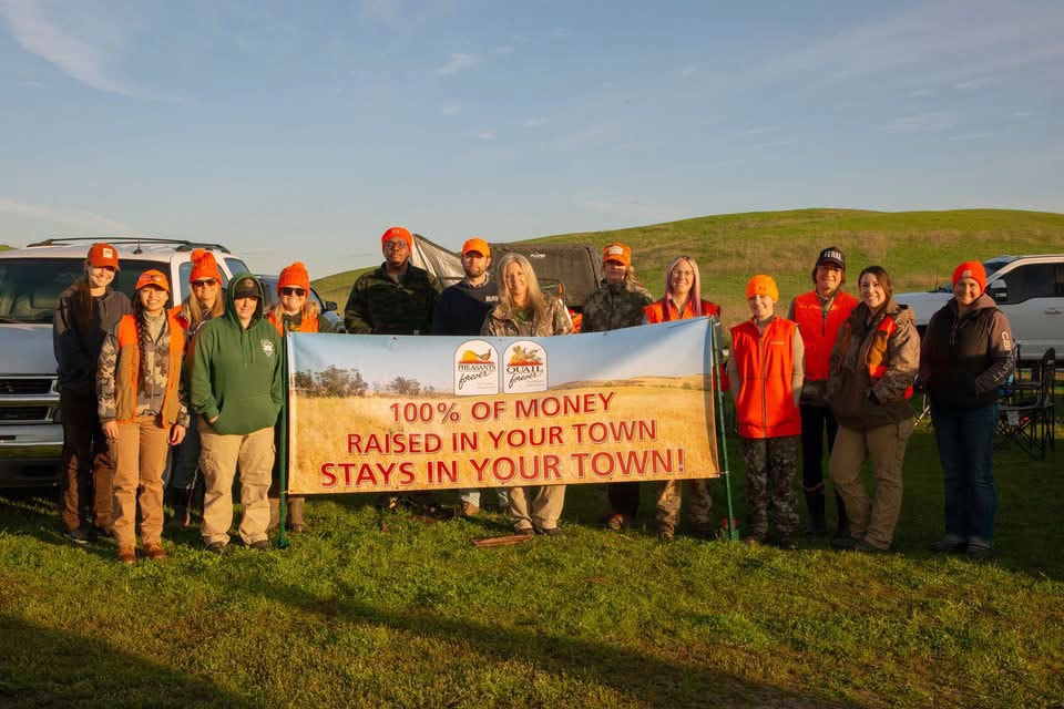 Large group of hunters in orange vests holding a Pheasants Forever banner reading '100% of money raised in your town stays in your town' at sunrise