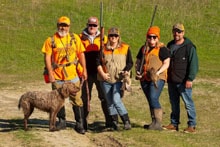 Group of five hunters in orange vests posing with a dog and harvested pheasants in a green field