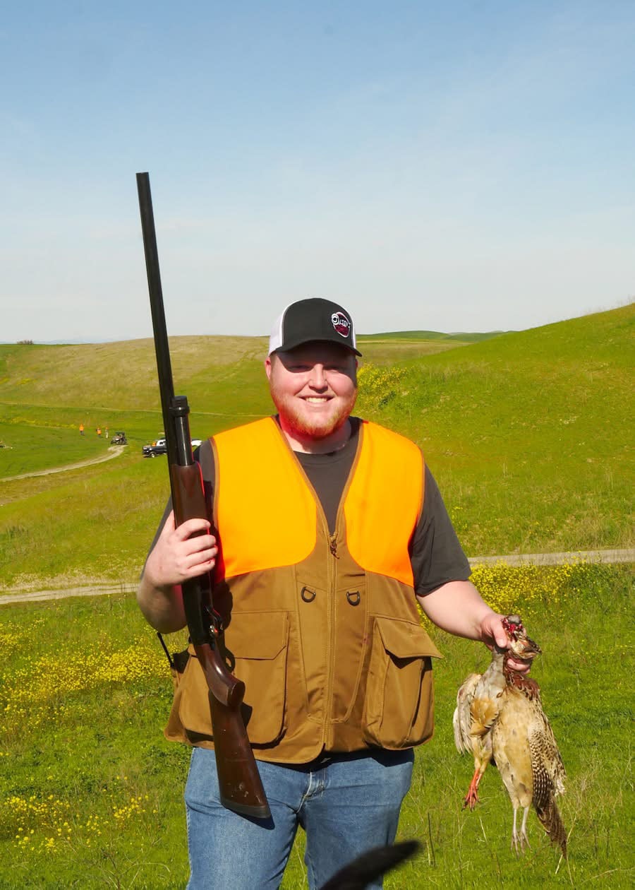 Smiling hunter in orange vest holding a shotgun and a harvested pheasant in a green hillside field