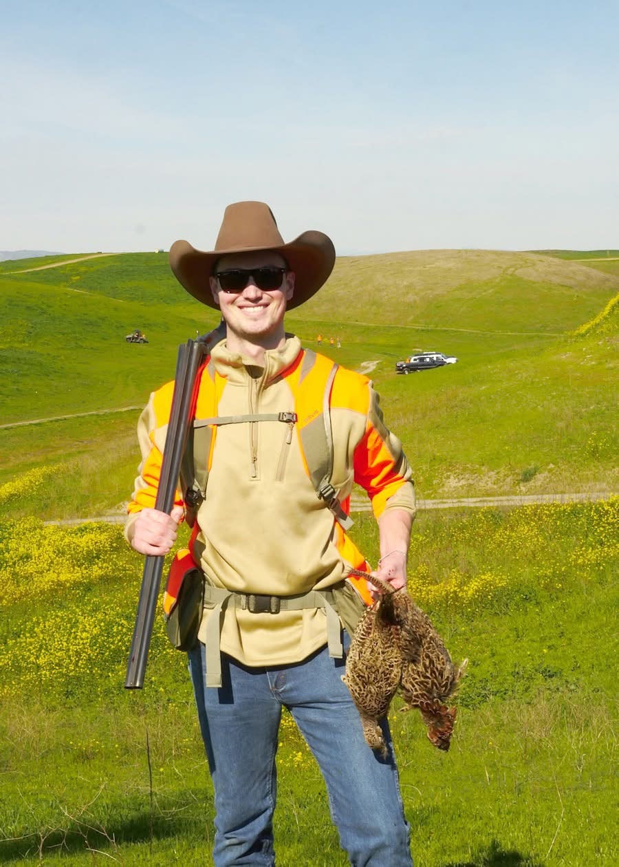 Hunter in orange vest and cowboy hat holding a shotgun and harvested pheasants on a green hillside with yellow flowers