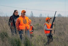 Mentor in blaze orange vest providing field instruction to youth hunters in a grassland at the 2025 Youth Mentor Hunt