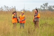 Three hunters in blaze orange vests — two youth and one adult — standing together in tall grass at the 2025 Youth Mentor Hunt